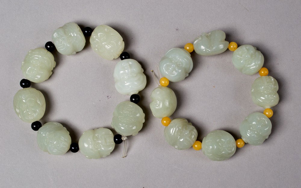 (2) Chinese Carved Jade Bracelets: Both eight beads, each one finely carved to depict a face, one separated by black stone beads, the other separated by yellow stone beads. Circa 20th century.