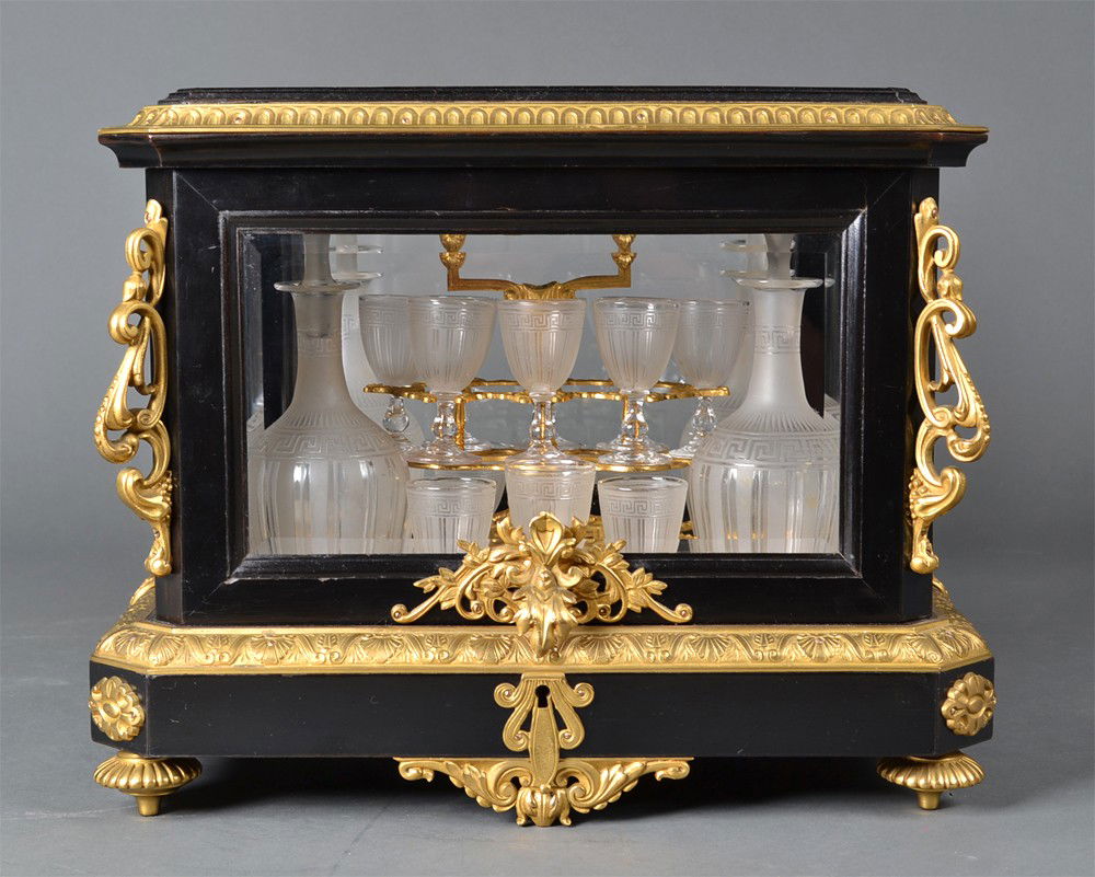 A Fine Gilt Bronze, Ebony Wood Cave a Liqueur: Having four Baccarat decanters and fourteen Baccarat cordials, decorated with Greek Key border and alternating stripes, housed within a dore bronze and ebony wood case. with beveled glass with egg and