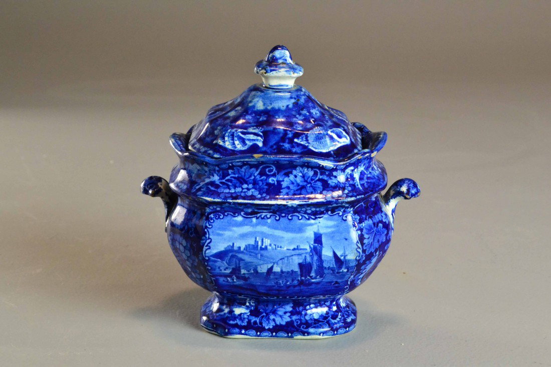 A Flow Blue Transferware Sugar Bowl: A two handled footed sugar bowl depicting castle and ship scenes on both sides with further leaf, grape and shell decorations. Impressed (wood and #29) to underside. Circa late 19th early 20th century