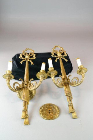 (2) BRASS ARM ELECTRIC WALL SCONCES: A pair of decorative brass arm wall sconces, topped with a brass ribbon, with decorative hanging drapes and two tassles, with scrolled arms, with candlestick resting on a ribbed bobeche, 23" H, circa 