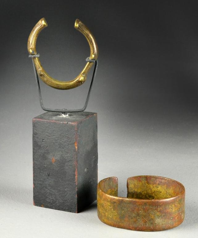 (2) African Tribal Copper Or Brass Cuff Bracelets: To include an unusual shaped cuff mounted in modern display stand, together with a very heavy possibly upper arm cuff with carved symbols, largest measures 3.5" D both very heavy, circa 19th century o