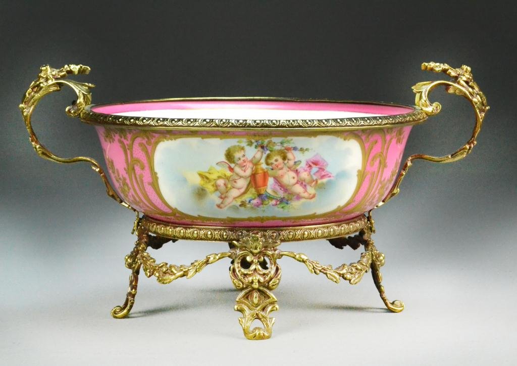 French Sevres Porcelain Bowl in Brass Mounts: Handpainted bowl has pink ground with gilded borders and image of playful cherubs, opposite side depicts floral design, white interior with floral border and central bouquet, ormolu handles and raised