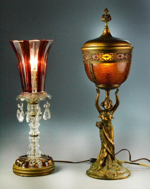 (2) Turn-of-the-Century Glass Lamps: Consisting of one bronze-painted metal lamp depicting a female figure holding up an amber glass covered urn housing light fixture along with a red & clear glass prism lamp with gold painted wooden cir