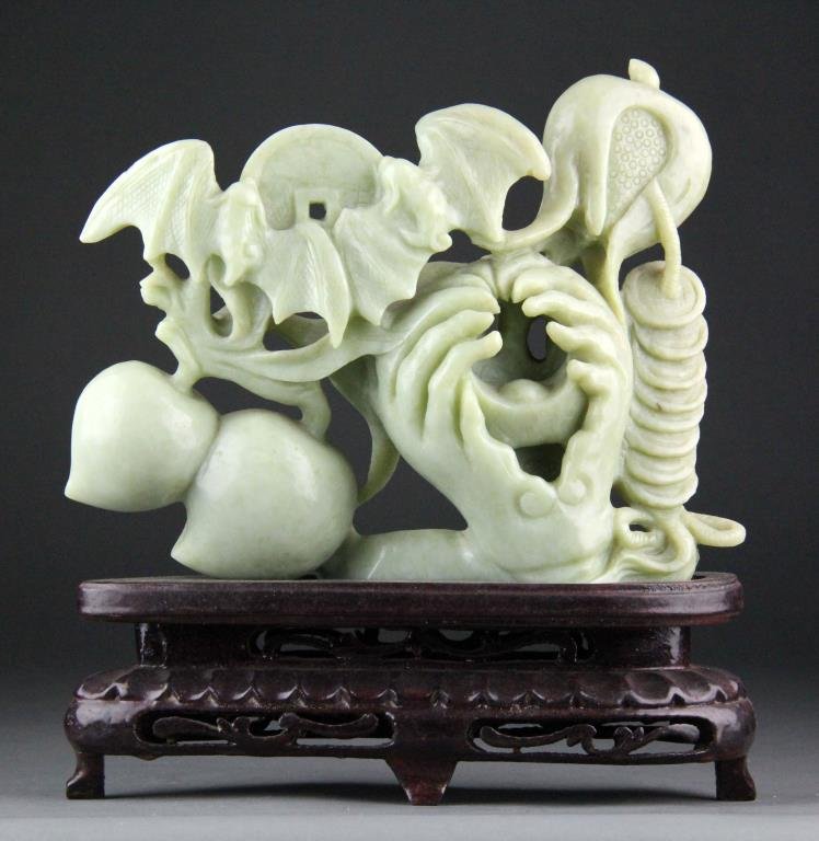 Chinese Jade Carving of Buddha Hand, Peaches & Bat: Finely carved jlight green jade depicting a coin ingot in Buddha's Hand, peaches, bats and many coins, along with carved wooden stand, 8" H x 9.5" L, circa 20th century, good condition.