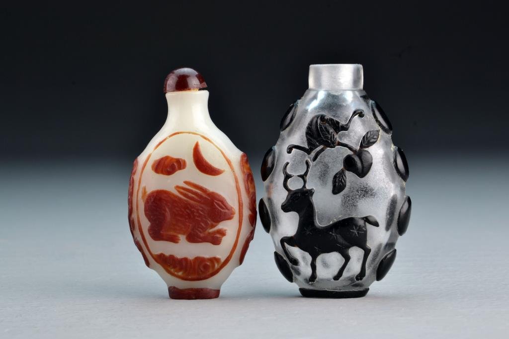 (2) Chinese Peking Glass Snuff Bottles: One having overlay depicting rabbit beneath moon and ruyi cloud on each side, having beast head handles, second example depicting deer and bat with peach, tallest is 2.9"H. From the collection of Davi