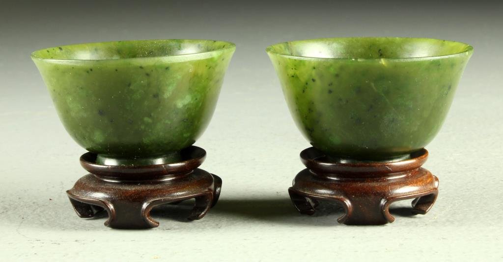 Pr. Chinese Qing Carved Jade Wine Cups: In spinach jade having black inclusions, each raised on short foot, each exhibiting a finely polished surface, both raised on hardwood stands, cup measures 1.25"H x 2.25"Dia., circa 19th century.