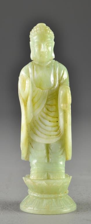 Chinese Carved Jade Figure of Buddha: Carved to depict a standing Buddha raised on double lotus base, the stone an even celedon with areas of green inclusions, 6"H. Condition: Chipping to base commensurate with age.