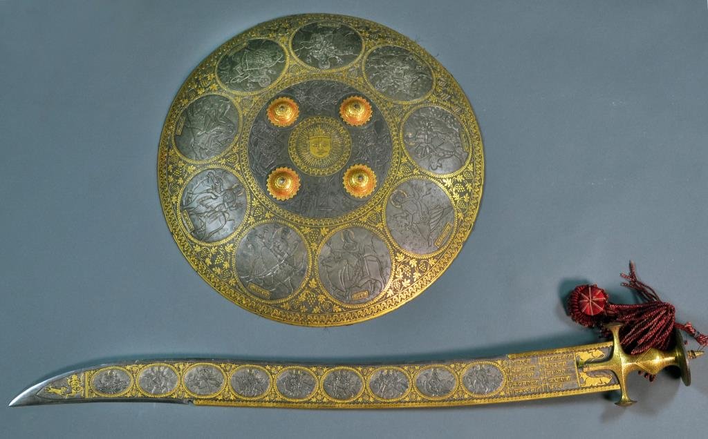 A Fine Indo-Persian Sword and Dahl Cuff Guard Shield: The sword decorated on both sides with nine oval scenes carved in high relief surrounded by raised gilt or gold floral and scroll decoration, the tip and base of the sword decorated in gold dogs and a