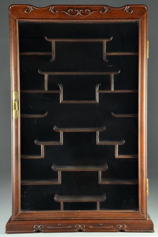A Chinese Rosewood Wall Hanging Cabinet: Having a shaped top with scroll decorations , glass panel door, the interior with green velvet backing, multiple shaped shelves and brass handle, 32"H x 21.5"W x 4.75"D, circa 20th century. Property o
