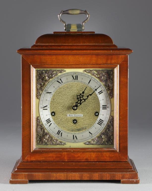 A Seth Thomas Shelf Clock: A carved wood shelf clock, possibly flame mahogany, having a metal swivel handle to top, push button glass door opens to the metal dial, framed by scrolling cherub decoration, having Roman numerals an