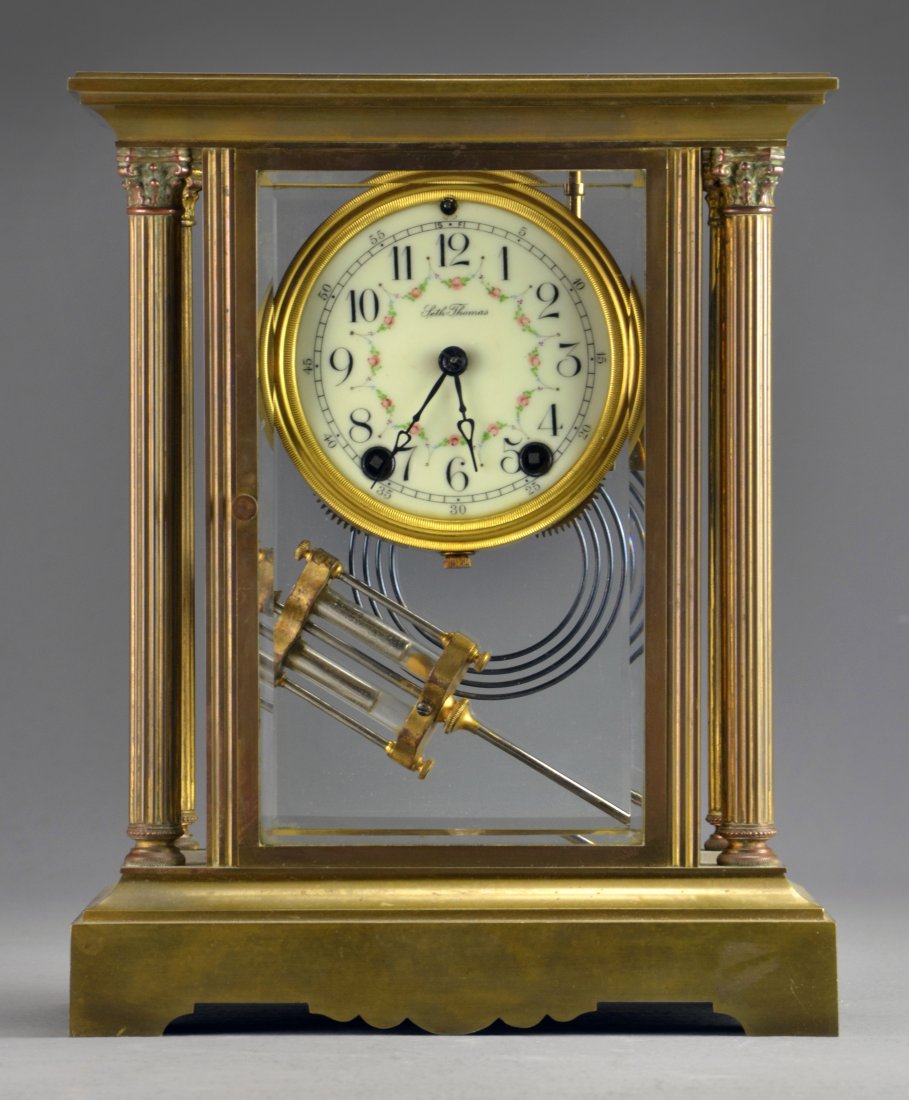 An Antique Seth Thomas Brass and Porcelain Mantel Clock: Having a brass case with four beveled glass panels and four corinthian columns housing a floral decorated porcelain clock face with faux mercury pendulum, measures 11.25"H x 9"W x 5"D, circa 1910. Con