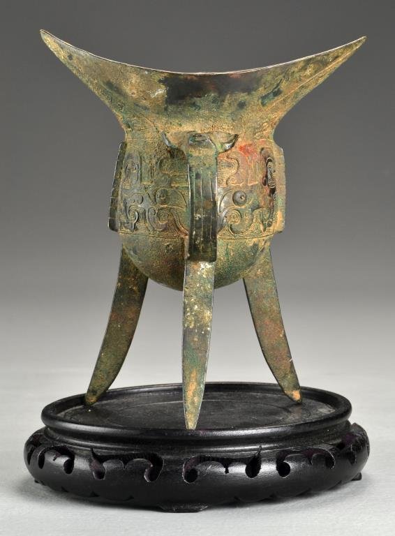 A Chinese Archaic Shang Style Bronze Jia Vessel: Having taotie mask separated by notched flanges, the handle adorned with a beast head in relief, having an inscription to the opposite side, raised atop tripod feet, now on a hardwood base, measures 8
