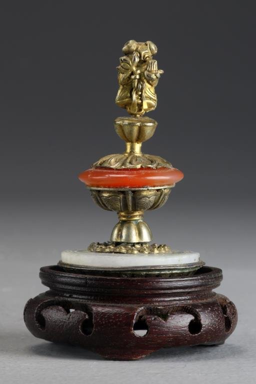 Chinese Qing Jade & Agate Hat Finial: Having a jade disc at bottom and an agate disc at center, further decorated with a figural brass figure atop lotus pad, the bottom having a molded Shou symbol, now displayed on a hardwood stand, 3.25"