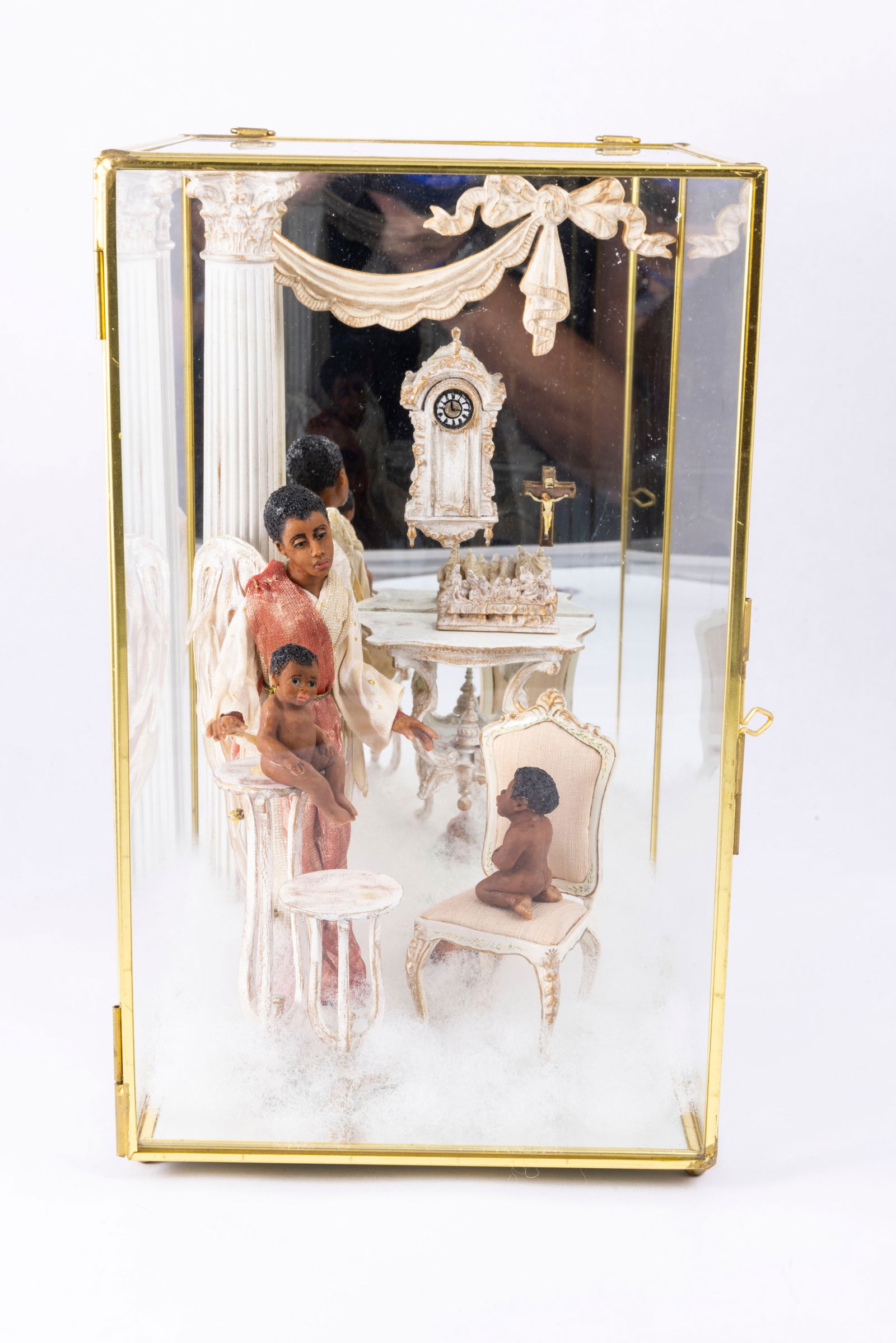 Hand Sculpted Male Angel Doll with Baby Children: A religious themed glass display with hand sculpted dolls. Two little babies in the nude with a male angel. There's Bespaq furniture with a chair and tables. There's a clock, a crucifix and more in th
