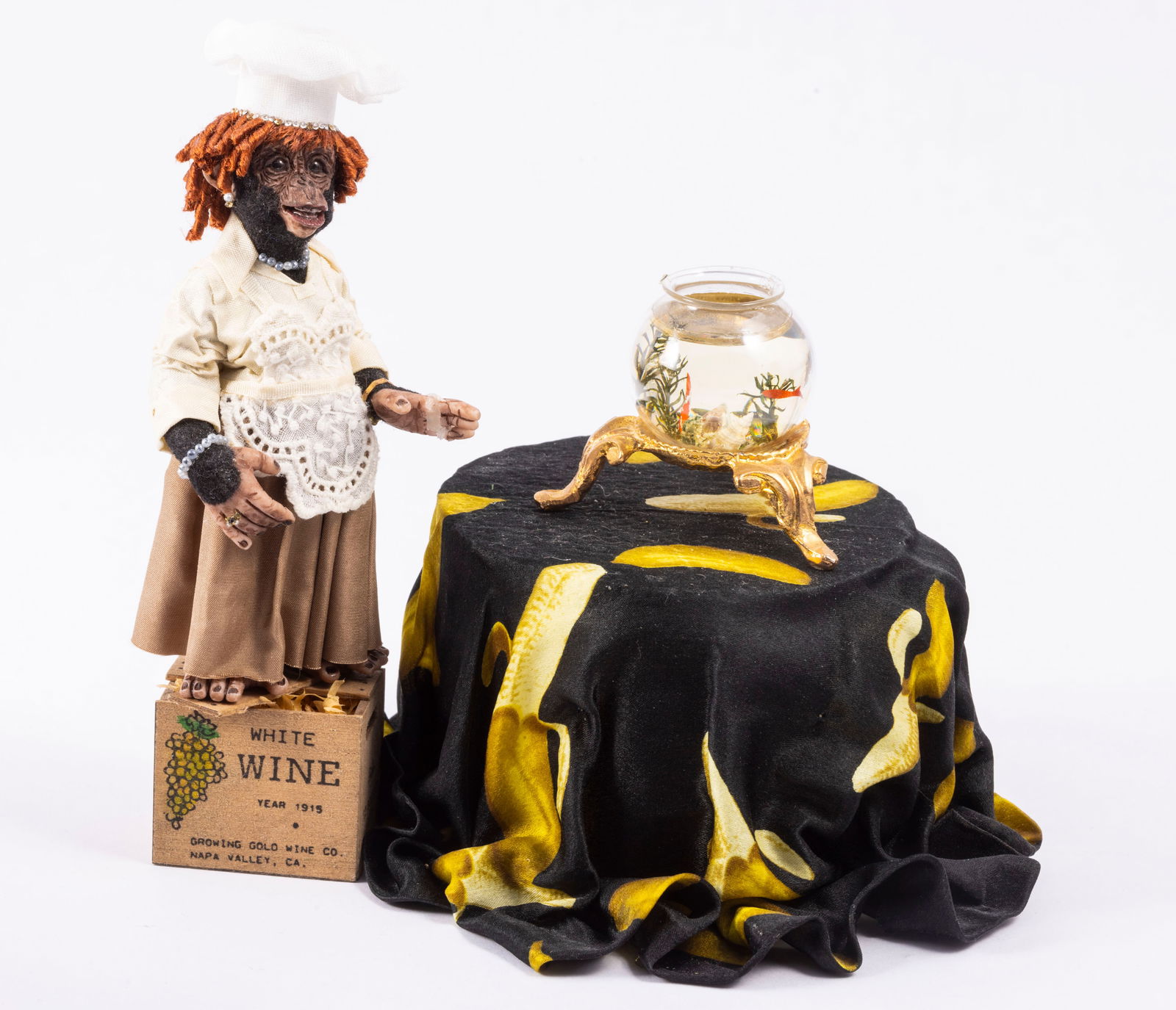Orangutan Girl in Dress Standing on Wine Box with Banana Table by Todd Krueger: Todd Krueger Orangutan in a dress with a white chef hat. She's standing on a wine box, and is missing a few fingers. There's a banana table next to her with a fish bowl on top. Please note her hand ha