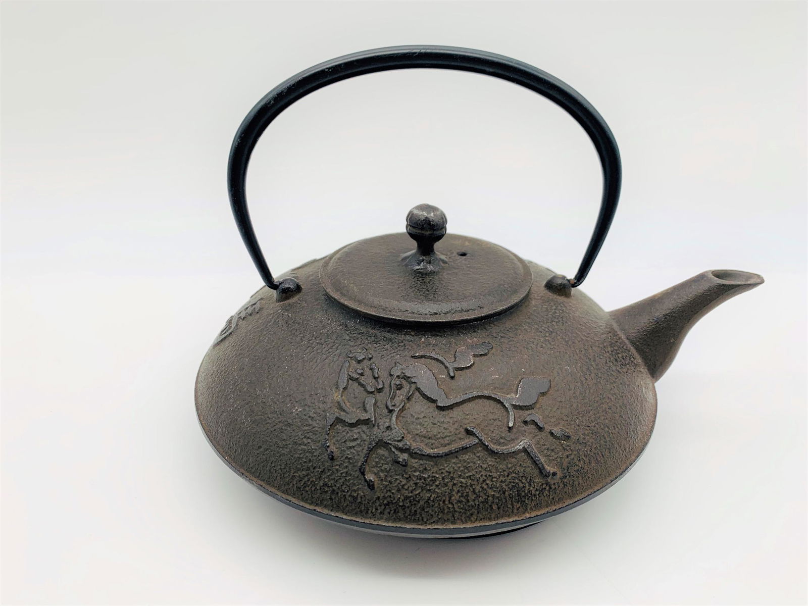 Joyce Chen Cast Iron Teapot: Joyce Chen Cast Iron Teapot. Decorated with running horses motif in relief. Includes mesh strainer and trivet. Signed. Measures 4" H not including handle x 9" W.