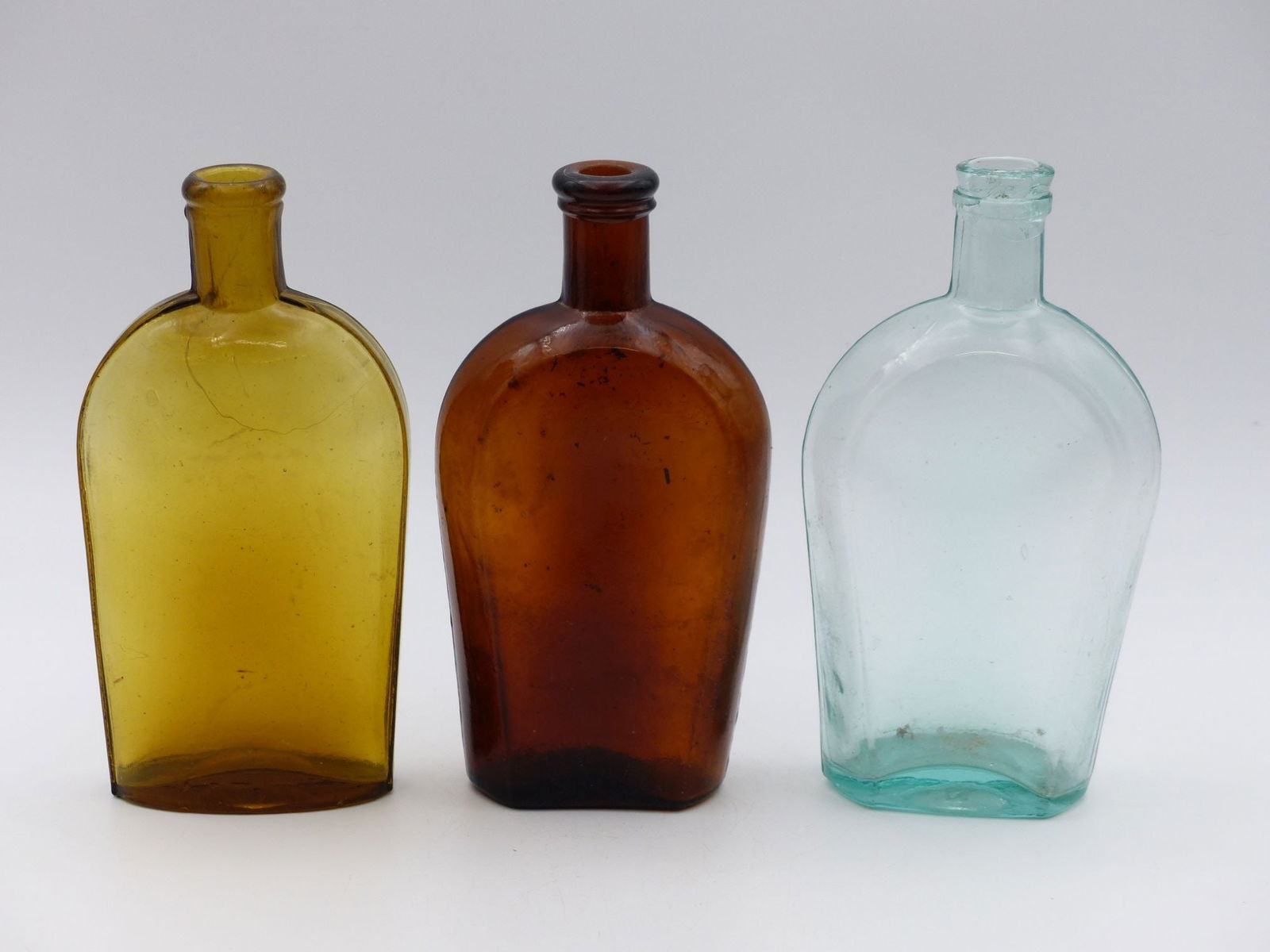 "Pint Flask Collection: Amber: This exquisite collection features three elegant pint flasks, each showcasing a unique amber hue. The flasks exhibit distinctive shapes and craftsmanship, characteristic of historical glass making, ma