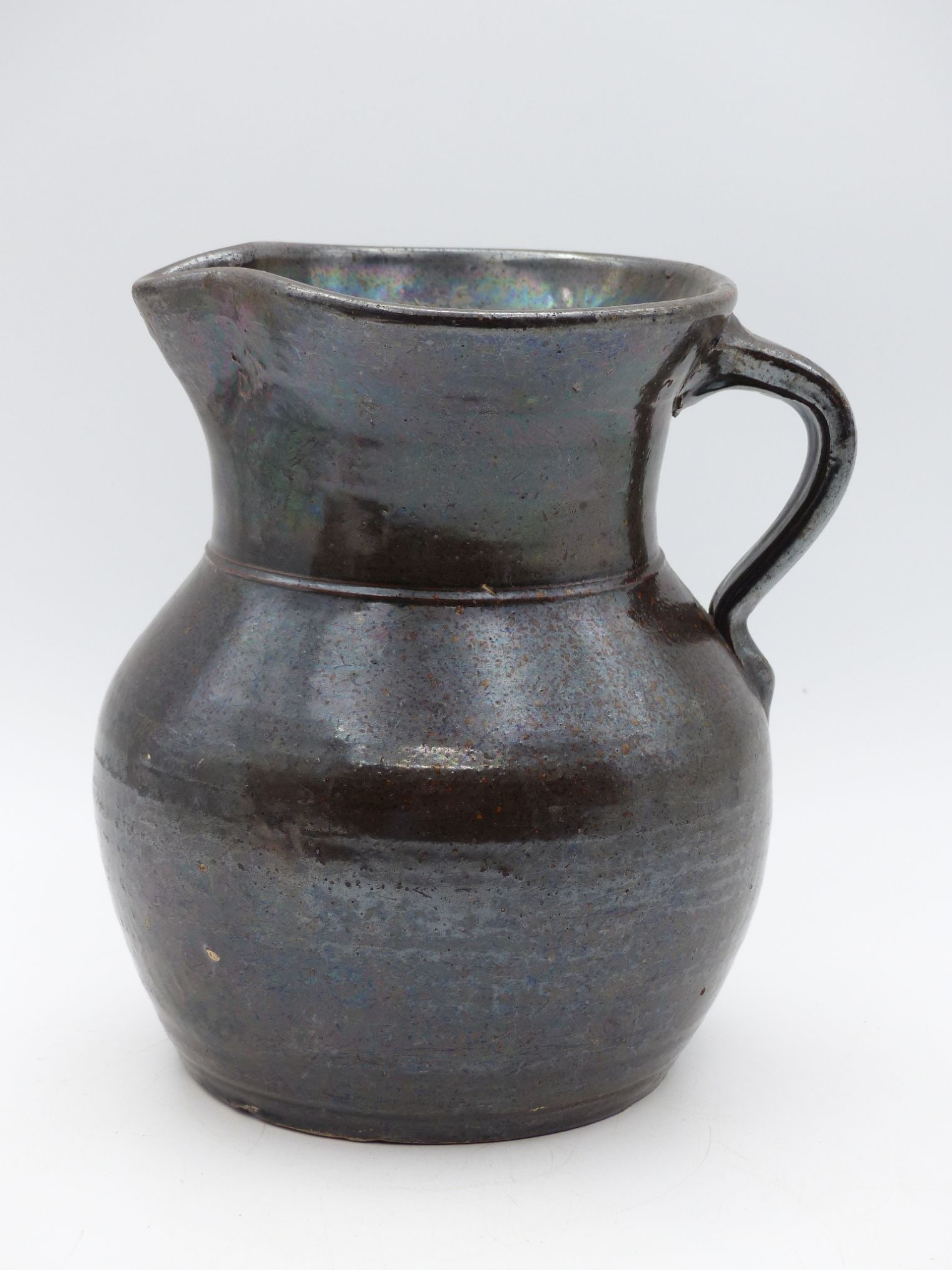 Albany Slip Pitcher (No Damage): This Albany slip pitcher showcases the classic beauty of utilitarian pottery, characterized by its rich, dark brown glaze with iridescent overtones. The gracefully flared spout and robust handle indic