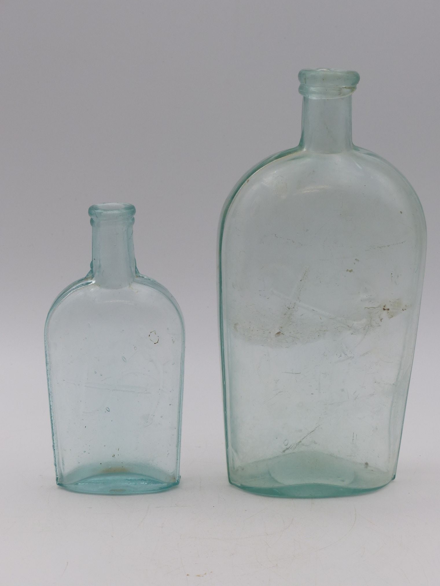 Aqua Flask Pair with Embossed Anchors: This exquisite pair of aqua flasks, featuring embossed anchors, exemplifies classic glass craftsmanship from the late 19th to early 20th century. The bottles showcase a beautiful gradient of aqua blue