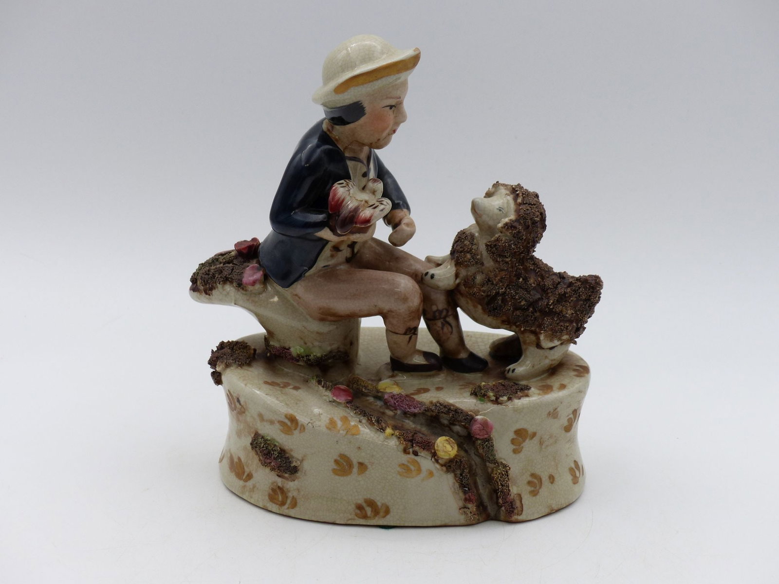 Ceramic Figurine of a Child with a Sheep: This delightful ceramic figurine depicts a child seated on a whimsical base, tenderly interacting with a sheep. The child is wearing a blue jacket and a straw hat, while the sheep features a textured,