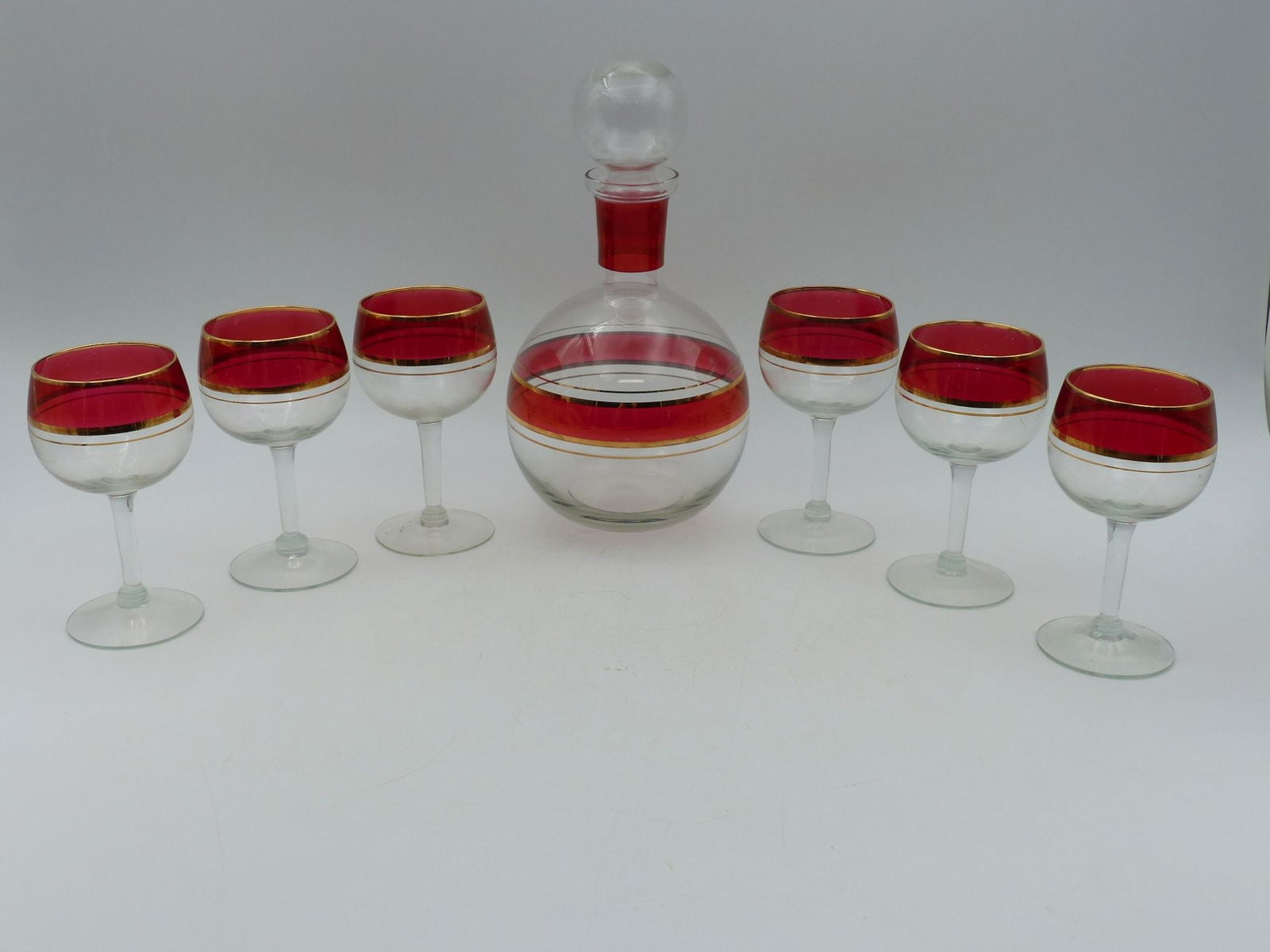Ruby & Gold Decanter Set with Six Wine Glasses: This elegant Ruby and Gold Decanter Set, accompanied by six exquisite wine glasses, harmonizes functionality with artistry. The decanter features a sophisticated spherical design with a defined gold t
