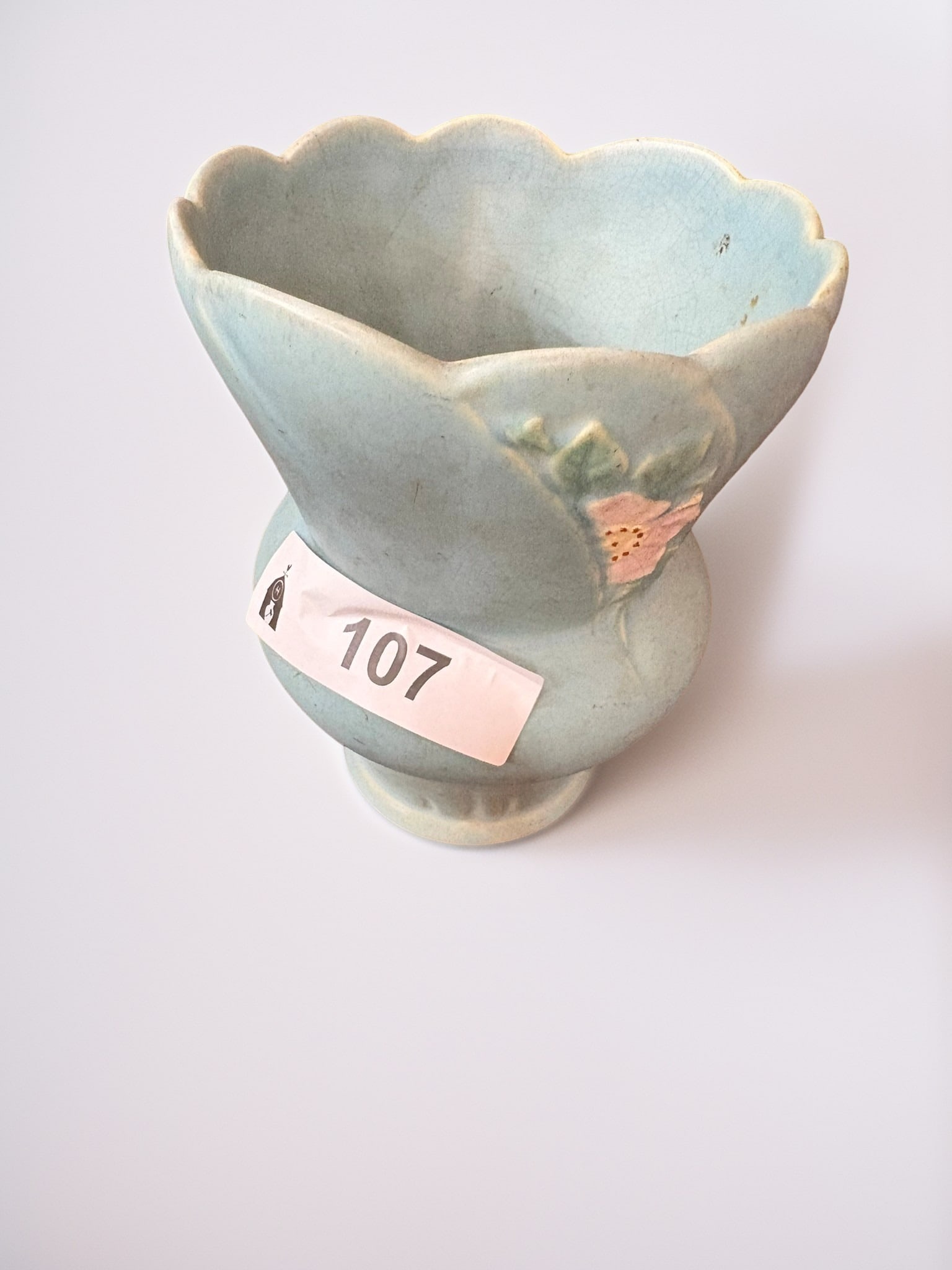 Weller Pottery Floral Vase: This elegant Weller Pottery vase features a soft, matte blue glaze and a charming molded floral relief detail. The piece is crafted with a wide, scalloped rim that tapers gracefully to a pedestal base