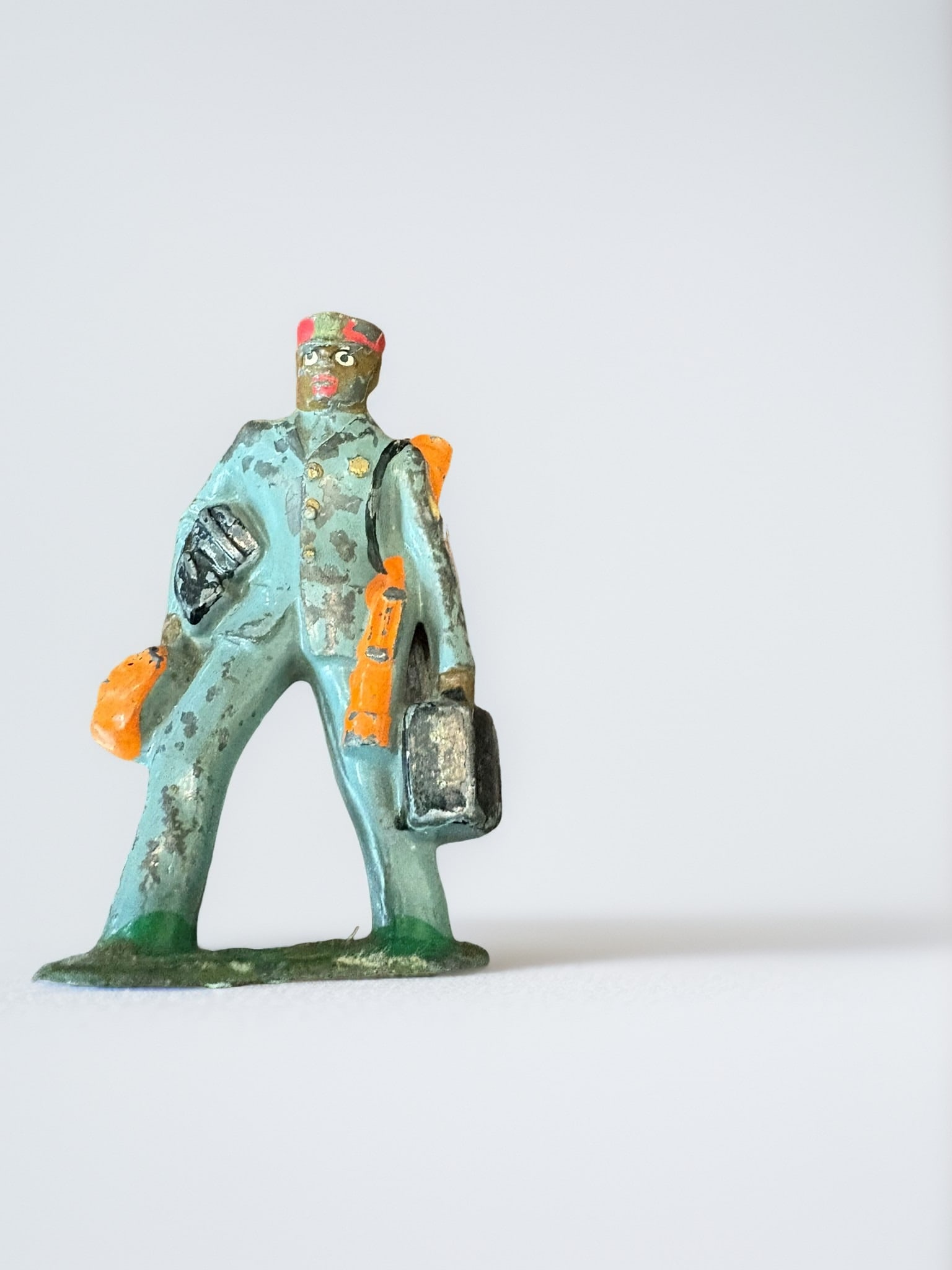 Vintage Barclay B-159 Train Porter Lead Figure: An original vintage lead toy figure manufactured by Barclay. This piece represents a train porter, painted in a blue uniform with orange accents, carrying luggage. It is a classic example of