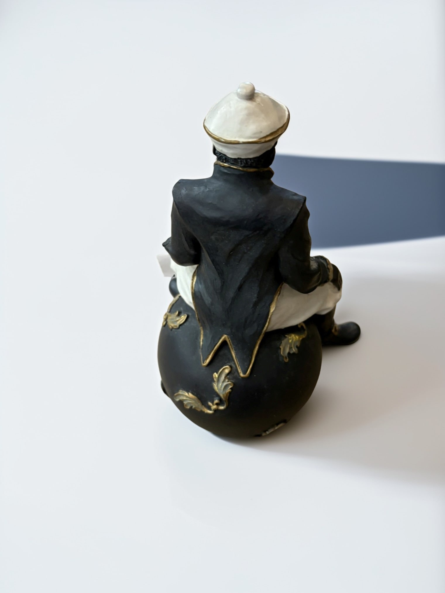Figural Ceramic Gentleman Sitting on Sphere: This decorative ceramic figurine features a dapper gentleman dressed in a formal tailcoat and cap, perched atop a spherical base. The piece is finished in a high-contrast palette of black, white, and 