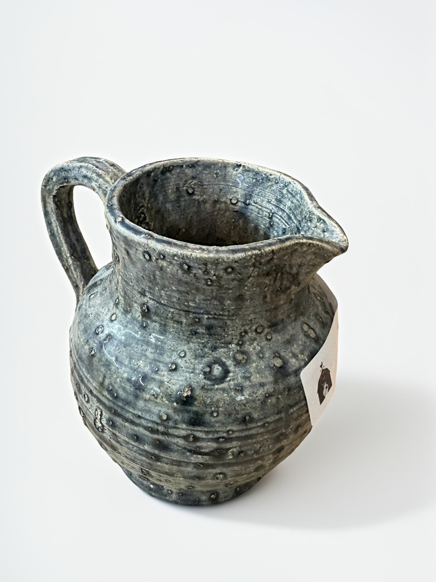Penny Cole Studio Pottery Pitcher, Greenwood, SC, 1976: A studio pottery pitcher hand-thrown by Penny Cole in Greenwood, South Carolina. The piece features a distinct, heavily textured, dimpled, and mottled dark blue and grey glaze. It is incised on the un