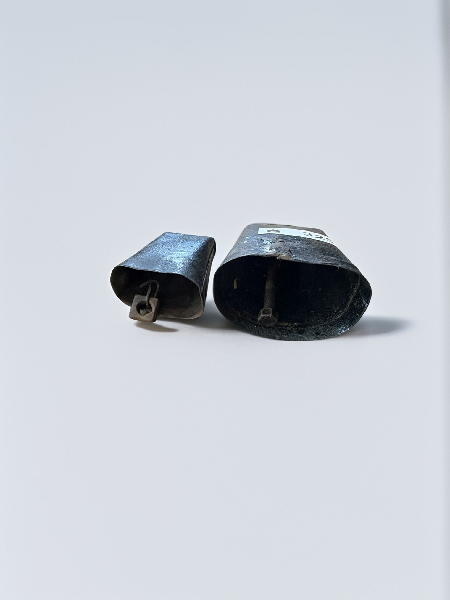 Set of Two Antique Farmstead Iron Cow Bells: This set features two rustic iron cow bells of varying sizes, exhibiting a weathered, dark patina consistent with historical agricultural use. Each bell retains its original metal clapper and carrying