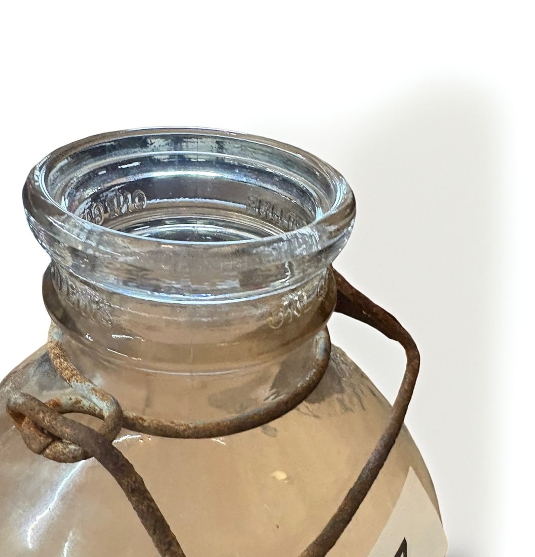 Vintage One-Gallon Clear Glass Dairy Milk Jug with Wire Bail Handle: This is a vintage, one-gallon clear glass milk jug, likely dating to the mid-20th century. It features a heavy-duty, rust-patinated wire bail handle attached to the neck for easy carrying. The collar
