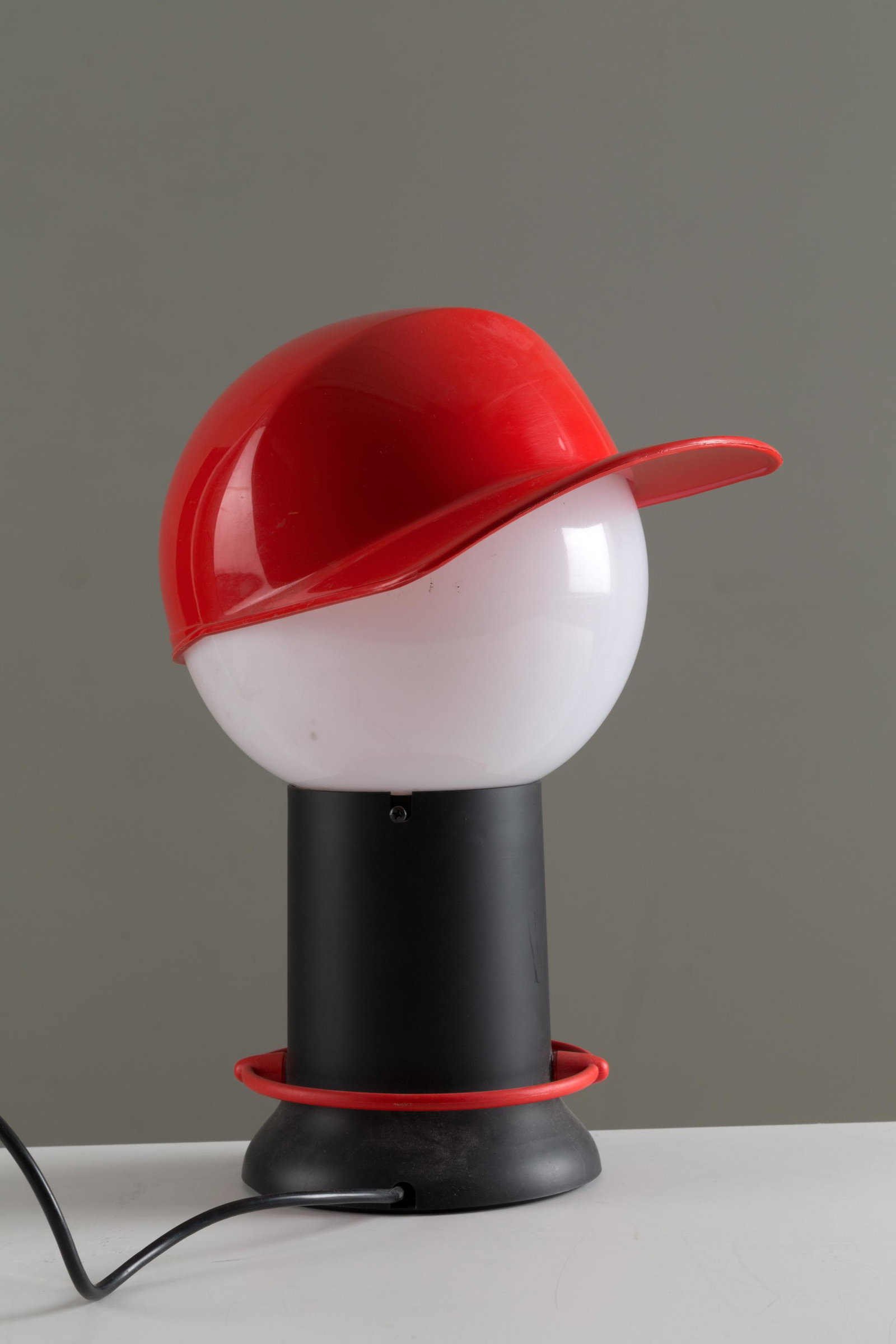 GIORGETTO GIUGIARO. Floor lamp mod. CAP. BILUMEN: GIORGETTO GIUGIARO (Garessio, 1938). Lampada da tavolo mod. CAP in plexiglass con cappello rosso. Produzione BILUMEN. Marchio del produttore sul fondo. Anni '70. Mis. Lung. cm. 30 Alt. cm. 36 ca. Val.