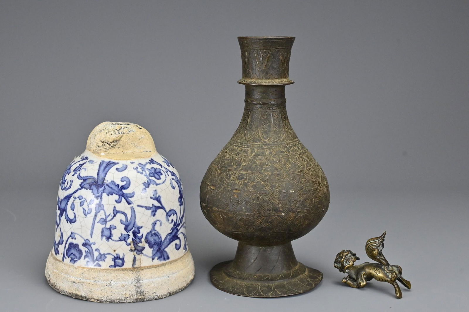 An Indo-Persian antique bronze vase on splayed foot: An Indo-Persian antique bronze vase on splayed foot decorated with a floral pattern together with a bell-form ceramic item with blue and white glaze of floral scrolls and a bronze figure of Foo dog. B