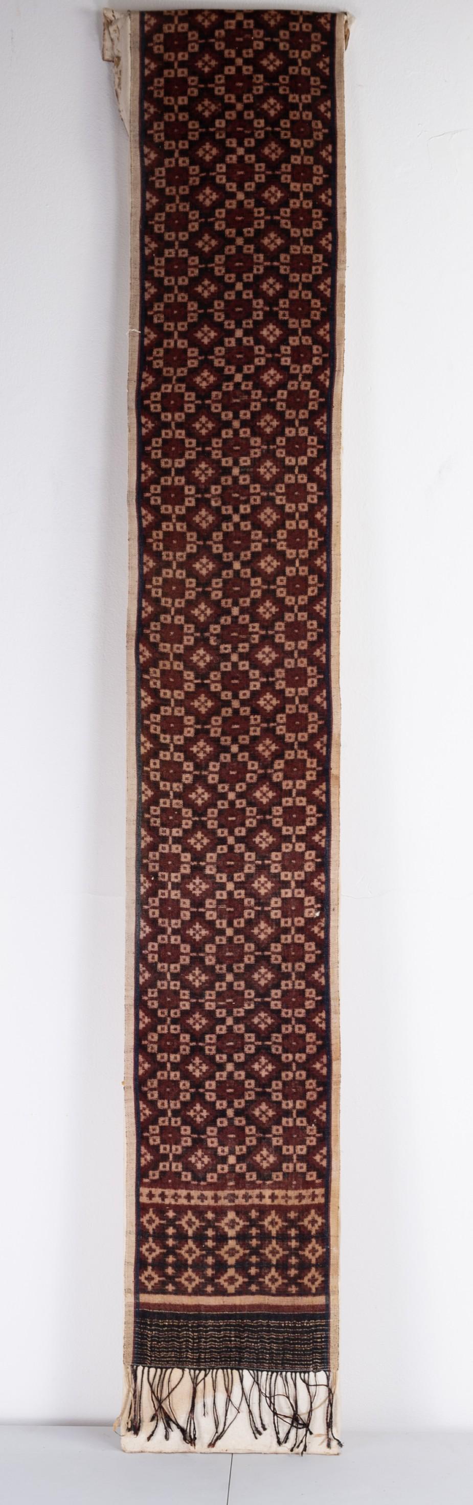 A BALINESE IKAT EMBROIDERY TEXTILE: A BALINESE IKAT EMBROIDERY TEXTILE. With geometric design in indigio and deep red with fringes to one end. Mounted loosely on board. 154cm length x 23cm wide. Condition: Fabric in good general co