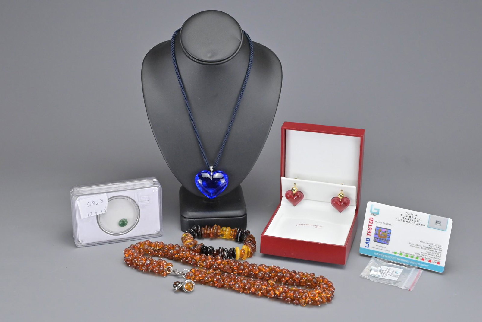 A GROUP OF JEWELLERY ITEMS AND LOOSE STONES: A GROUP OF JEWELLERY ITEMS AND LOOSE STONES. To include a boxed Baccarat blue crystal heart pendant with silver mount and clasp together with a pair of red crystal heart earrings. With amber bead neck