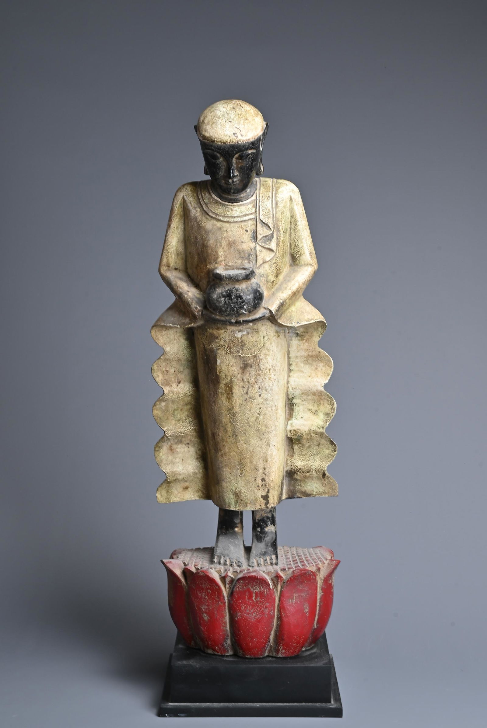A BURMESE CARVED WOOD FIGURE OF A MONK. The figure standing dressed in robes holding a bowl on a: A BURMESE CARVED WOOD FIGURE OF A MONK. The figure standing dressed in robes holding a bowl on a lotus base. Mounted on a later display stand. 62cm tall.