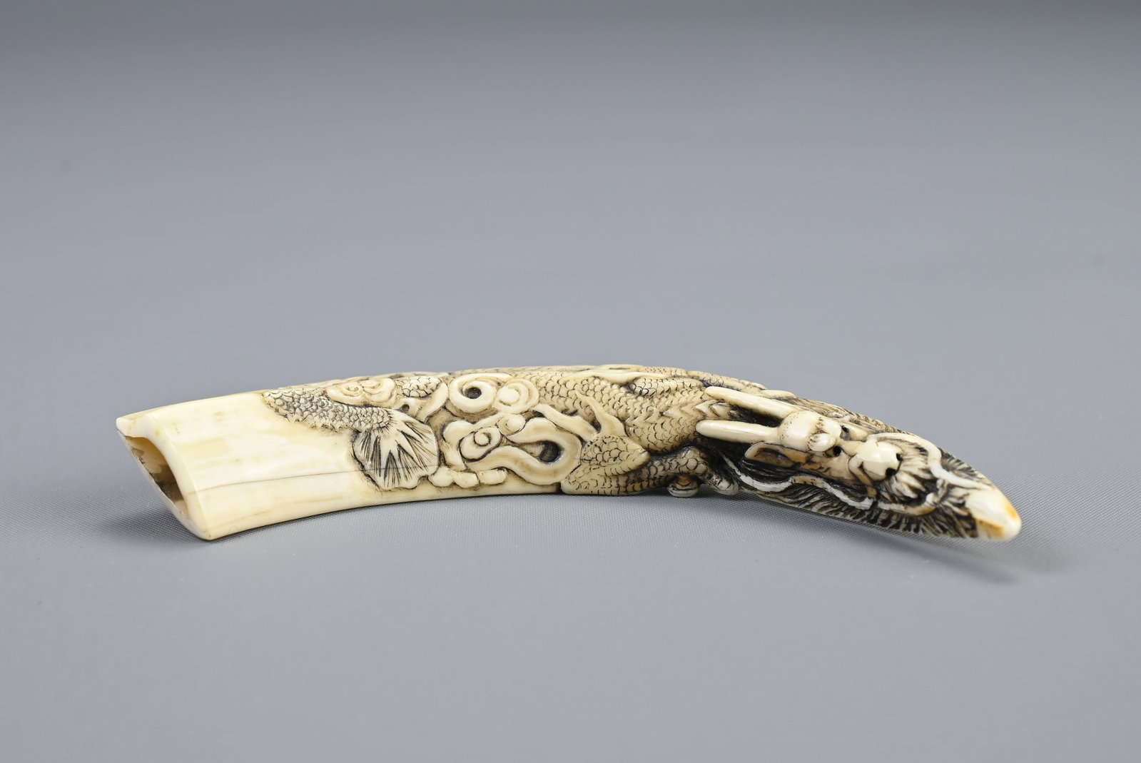 JAPANESE SHOWA OR HEISEI PERIOD IWAMI BOAR TUSK NETSUKE, 20th century, Carved to one side with a: JAPANESE SHOWA OR HEISEI PERIOD IWAMI BOAR TUSK NETSUKE, 20th century, Carved to one side with a scaly dragon emerging from scrolling clouds, the reverse with a short sword the beast holds in its claw