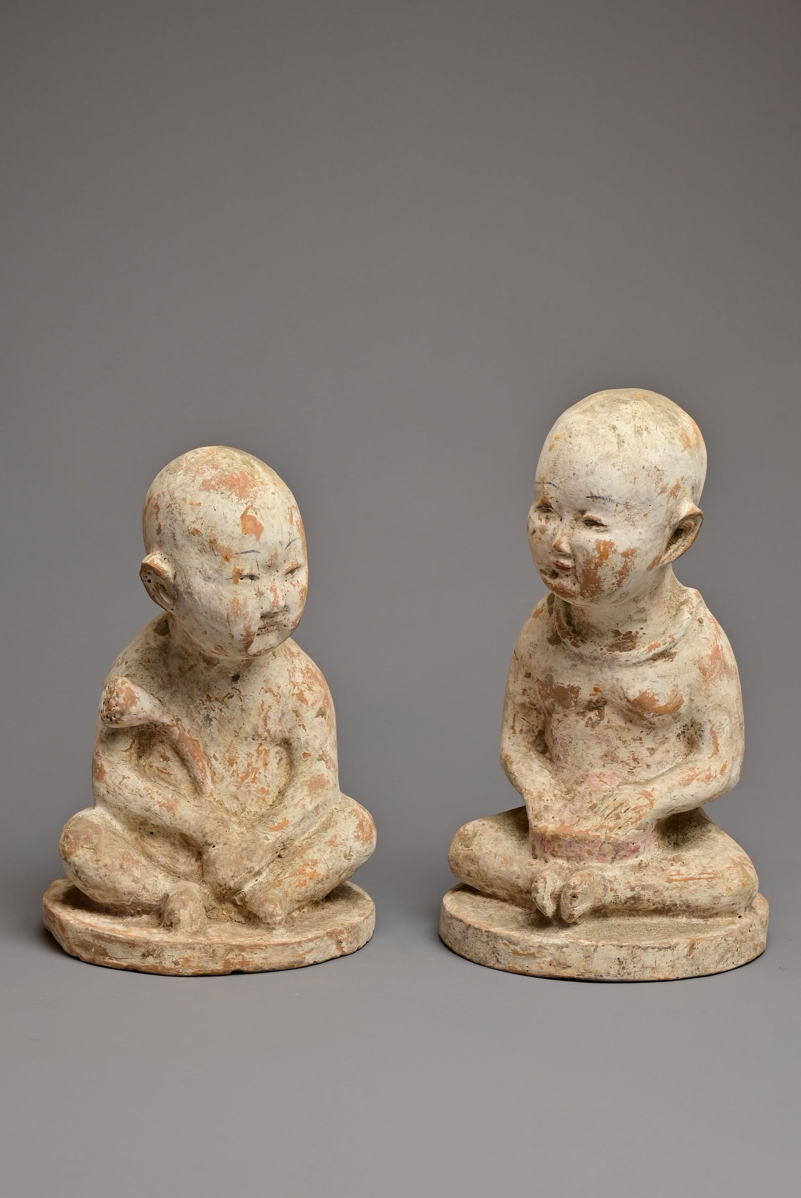 A PAIR OF CHINESE PAINTED POTTERY FIGURES OF SEATED BOYS. Finely modelled figures of boys sitting: A PAIR OF CHINESE PAINTED POTTERY FIGURES OF SEATED BOYS. Finely modelled figures of boys sitting nude, one holding a lotus stem the other with a drum decorated in brown, red and black pigments natura