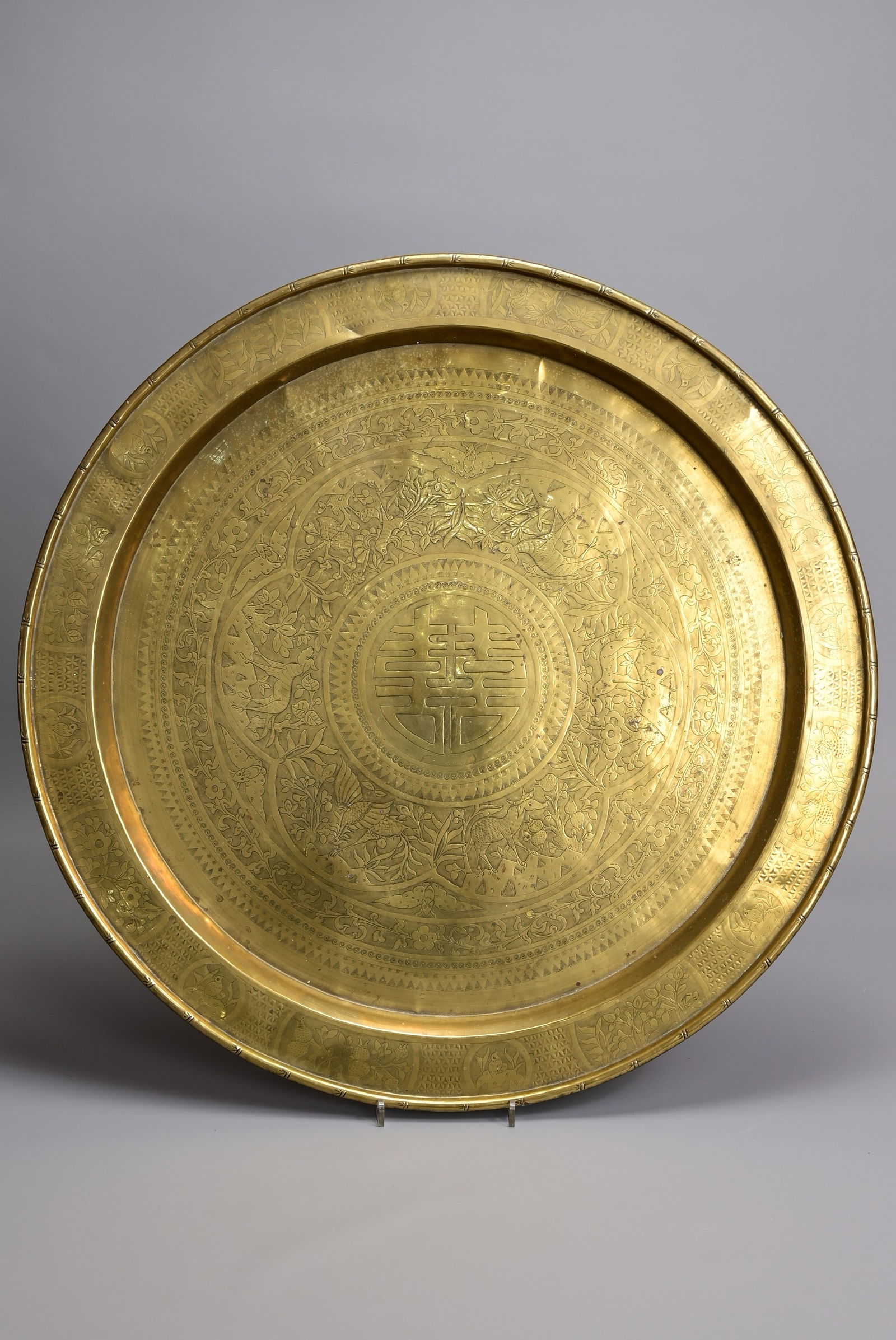 A 20TH CENTURY CHINESE ENGRAVED BRASS CIRCULAR TRAY TABLE TOP ON TURNED FOLDING WOODEN STAND. The: A 20TH CENTURY CHINESE ENGRAVED BRASS CIRCULAR TRAY TABLE TOP ON TURNED FOLDING WOODEN STAND. The circular brass tray engraved with a central Shou character, within a circular stiff-leaf edged cartouc