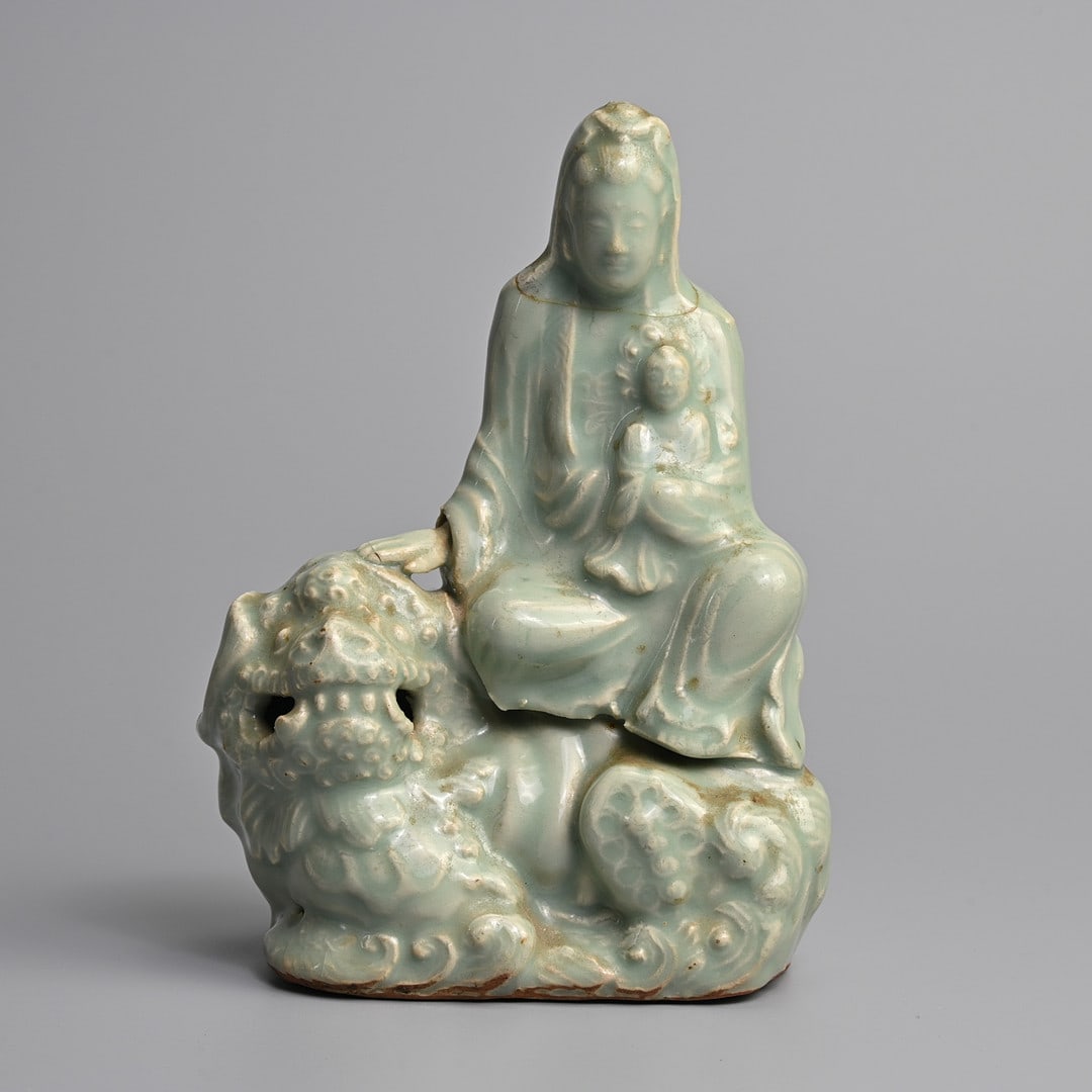 A CHINESE LONGQUAN CELADON GLAZED PORCELAIN FIGURE OF GUANYIN, MING DYNASTY (1368-1644).: Dressed in robes holding a child, seated on a Buddhist lion with her hand gently resting on its head. Covered overall in a pale celadon glaze. 19.5cm tall.
