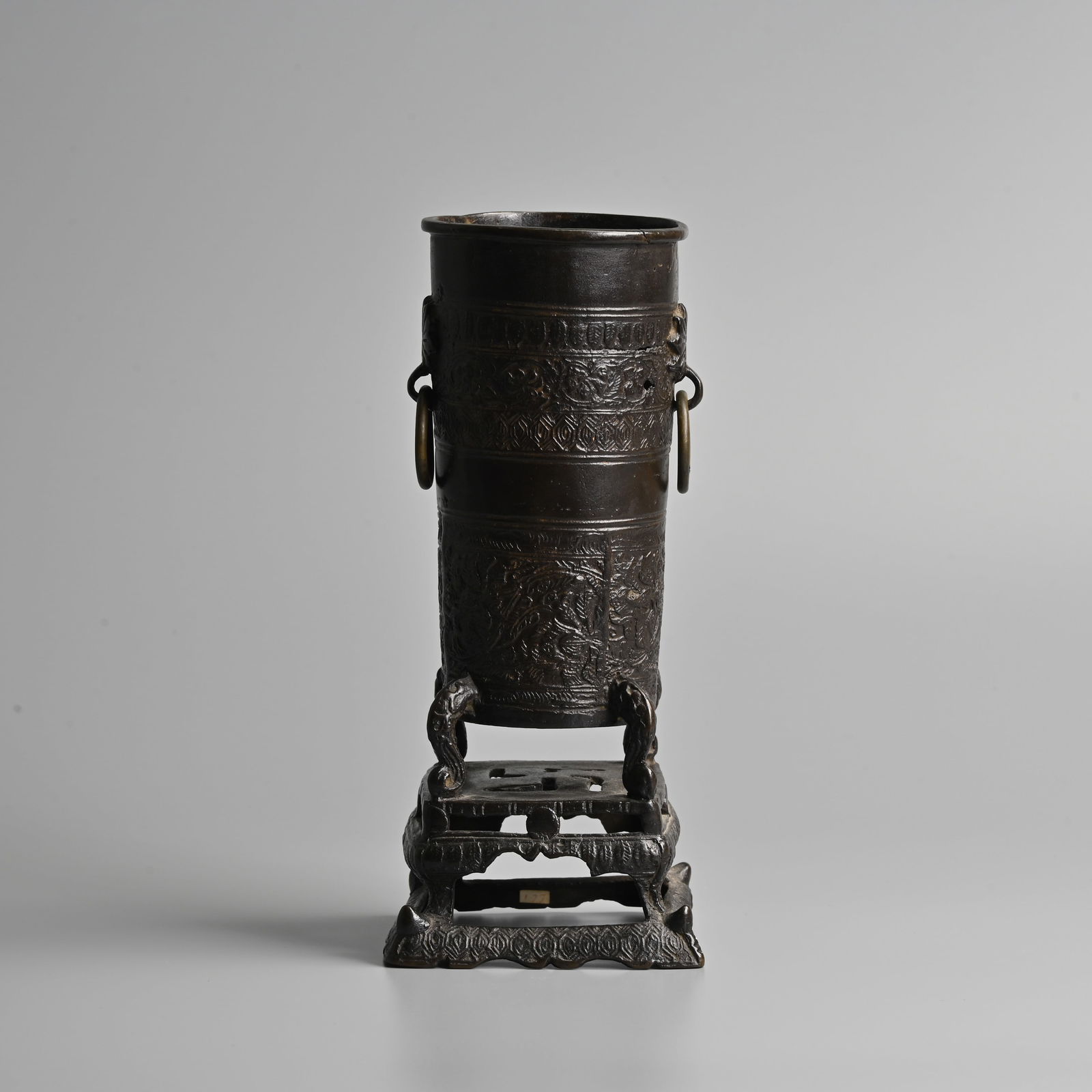 A CHINESE BRONZE BUDDHIST INCENSE STICK HOLDER, MING DYNASTY.: A CHINESE BRONZE BUDDHIST INCENSE STICK HOLDER, MING DYNASTY. Cylindrical pot decorated in relief with bands of floral scrolls, suspended ring handles raised on four scroll feet on an openwork stand w