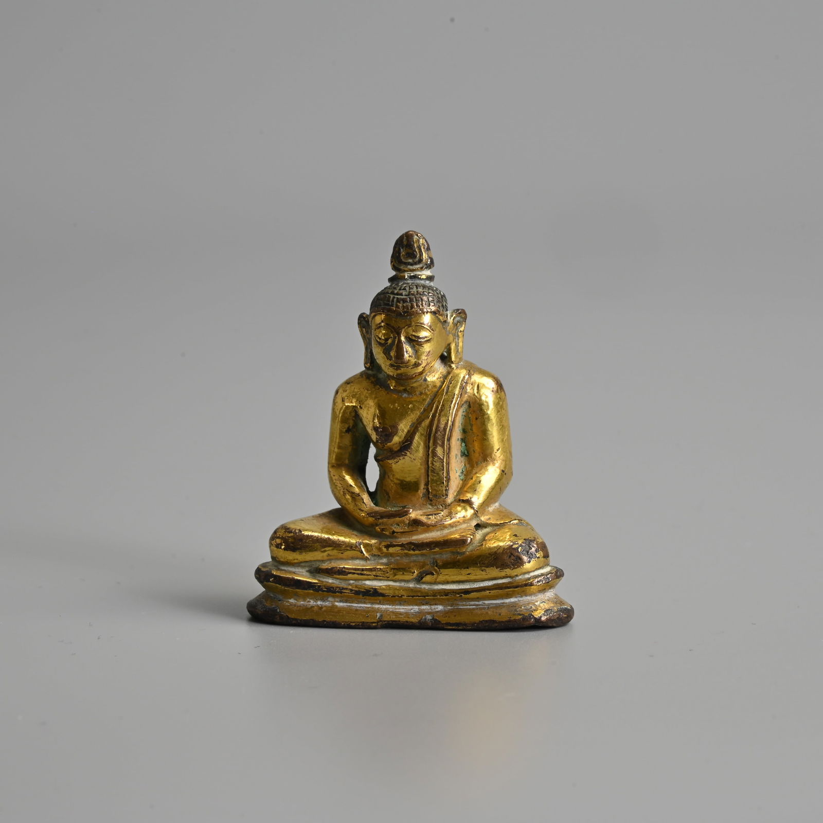 A SMALL GILT BRONZE SEATED BUDDHA, PROBABLY SRI LANKA, ANURADHAPURA STYLE, 18TH CENTURY OR EARLIER: A SMALL GILT BRONZE SEATED BUDDHA, PROBABLY SRI LANKA, ANURADHAPURA STYLE, 18TH CENTURY OR EARLIER Figure of Shakyamuni in meditation, dressed in robes with flames above the head. 5.2cm tall. Weight 8