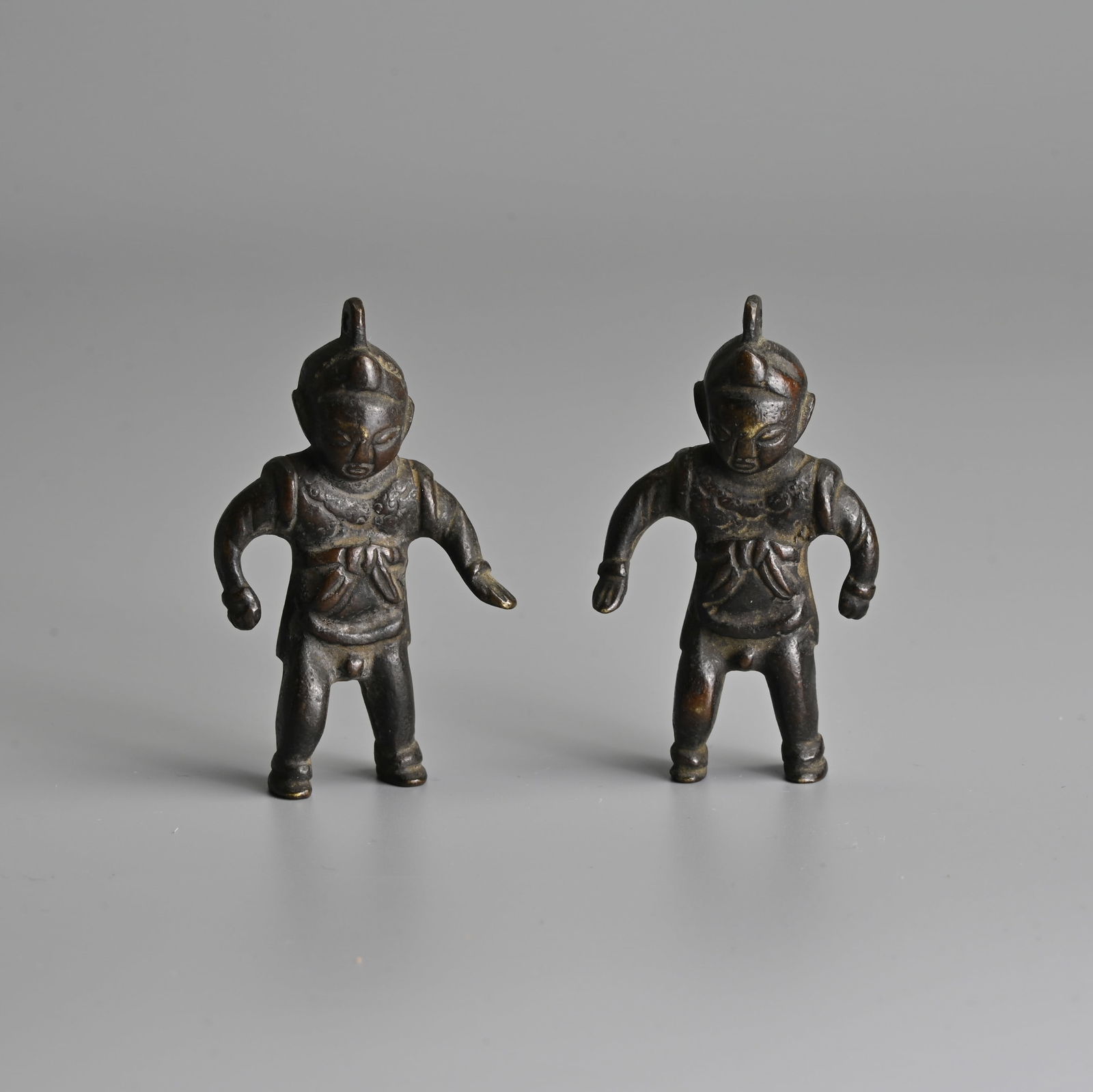 A PAIR OF SOUTHEAST ASIAN BRONZE DEITY PENDANTS, 19TH CENTURY OR EARLIER.: A PAIR OF SOUTHEAST ASIAN BRONZE DEITY PENDANTS, 19TH CENTURY OR EARLIER. Figures of boys wearing tunics tied at the chest. 6.5cm tall. (2). Condition: Both in good overall condition. *This lot is to