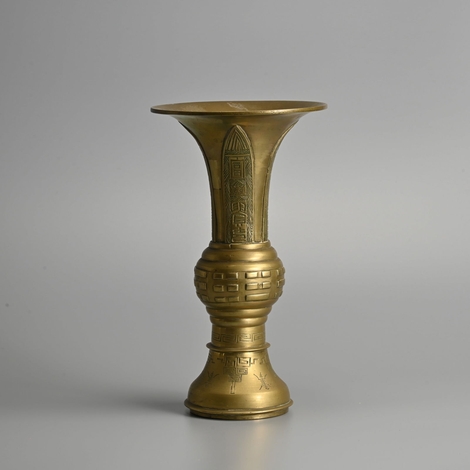 A CHINESE BRASS GU FORM VASE, LATE QING DYNASTY.: A CHINESE BRASS GU FORM VASE, LATE QING DYNASTY. The upper section relief decorated with quatrefoil stylised lotus leaves featuring mythical beast masks and characters of Fu, Gui, Shou, Kao. The middl