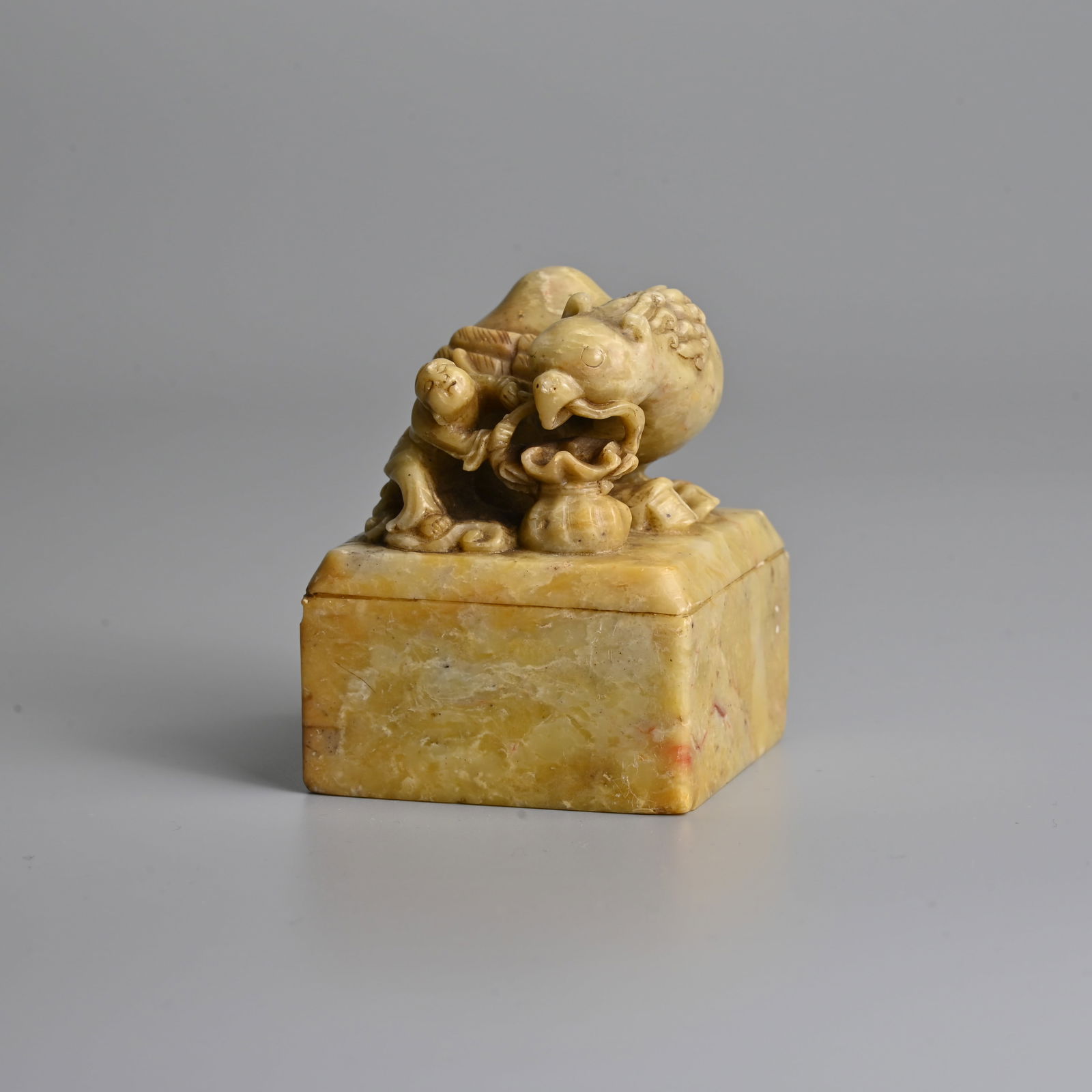 A CHINESE CARVED SOAPSTONE 'PHOENIX AND BOY' SEAL, 19/20TH CENTURY.: A CHINESE CARVED SOAPSTONE 'PHOENIX AND BOY' SEAL, 19/20TH CENTURY. Of square form with bevelled edge surmounted by a large phoenix with a tied bag in its beak with a boy holding onto the handle. With