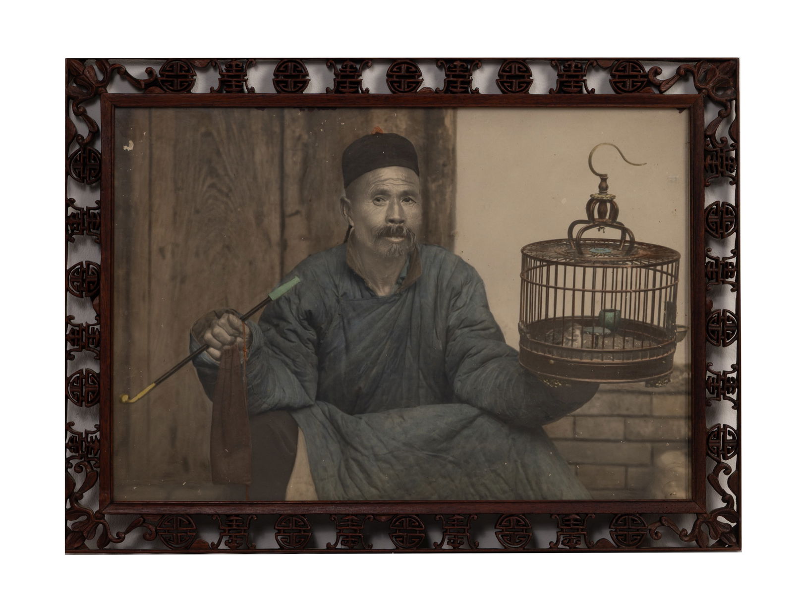 A FRAMED HAND COLOURED PRINT OF A CHINESE MAN, 20TH CENTURY.: A FRAMED HAND COLOURED PRINT OF A CHINESE MAN, 20TH CENTURY. Depicting a man seated holding a bird cage and pipe. Mounted in a carved and pierced 'Shou' wood frame and glazed. Total frame size 43.5cm