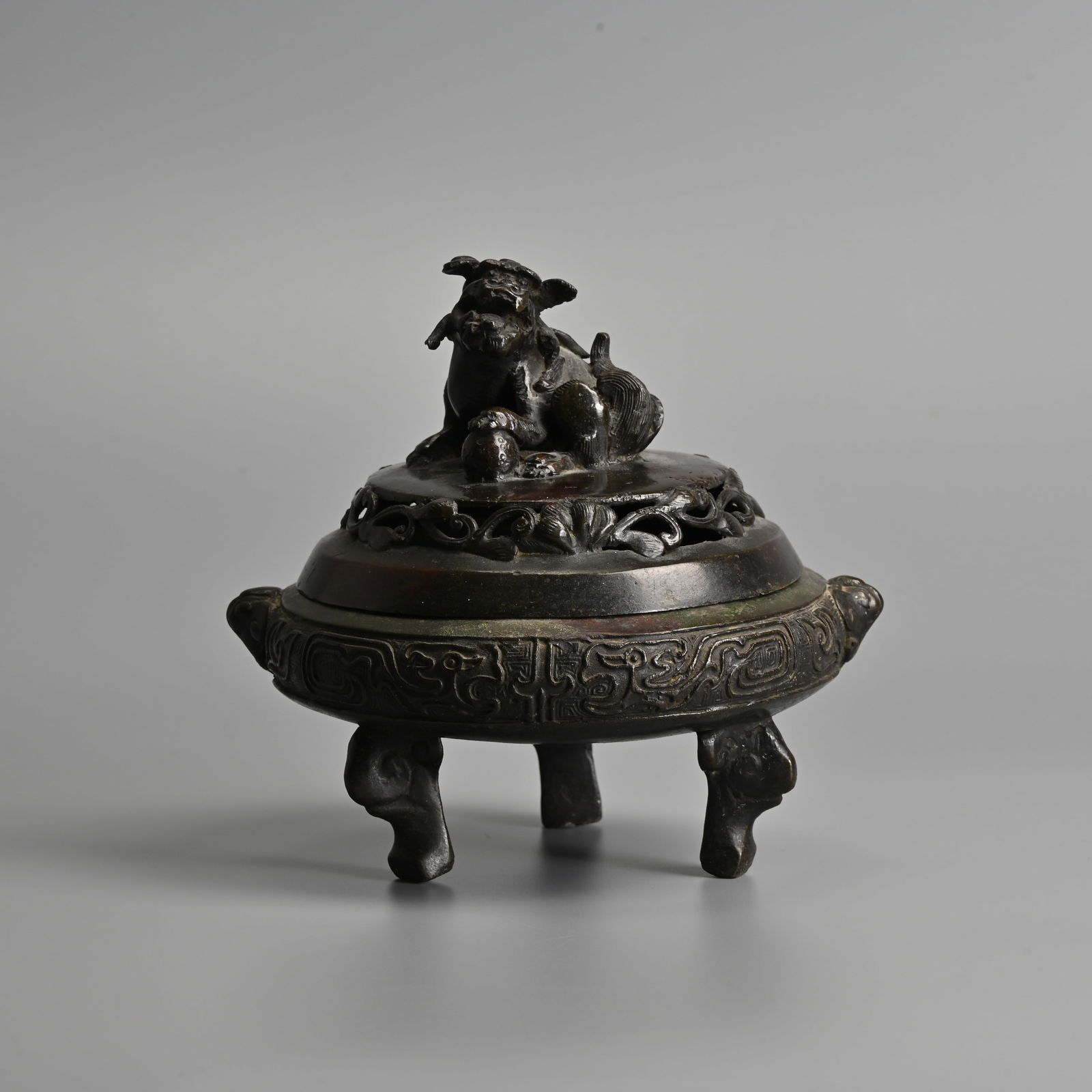 A JAPANESE BRONZE TRIPOD CENSER, MEIJI PERIOD.: A JAPANESE BRONZE TRIPOD CENSER, MEIJI PERIOD. Of squat form decorated in low relief with stylised archaic dragons and mask handles, raised on the animal form feet. The cover with pierced leafy scroll