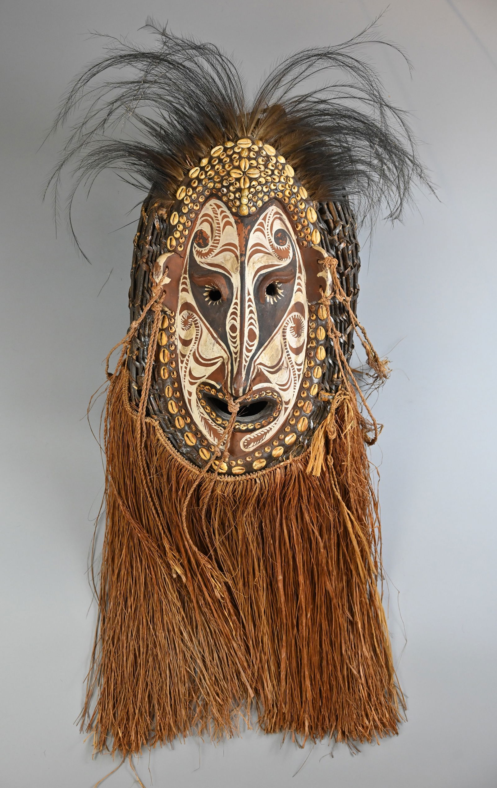 A LARGE PAPUAN GULF DANCE MASK, 20TH CENTURY, PROBABLY SEPIK RIVER REGION, PAPUA NEW GUINEA.: A LARGE PAPUAN GULF DANCE MASK, 20TH CENTURY, PROBABLY SEPIK RIVER REGION, PAPUA NEW GUINEA. The elongated painted wooden mask inset with cowrie shells, suspended woven reads and with feather headdres