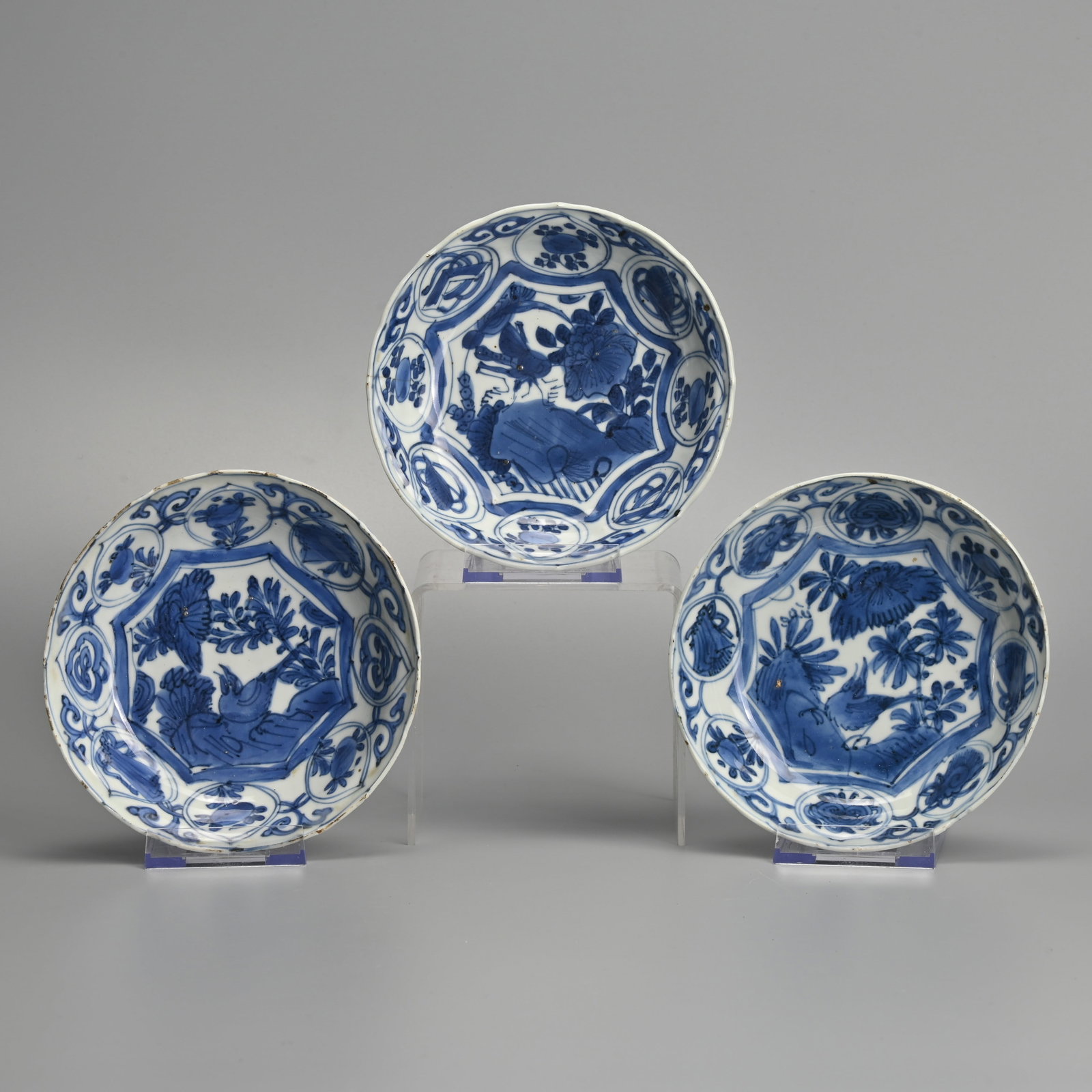 A MATCHED SET OF THREE CHINESE PORCELAIN BLUE AND WHITE SAUCERS, LATE MING DYNASTY, WANLI PERIOD: Each painted with a bird perched on rockwork before peony in landscape, the border with scrolls, flowers and fans within circular swagged cartouches, the undersides with foliate panels, approx.. 14 cm