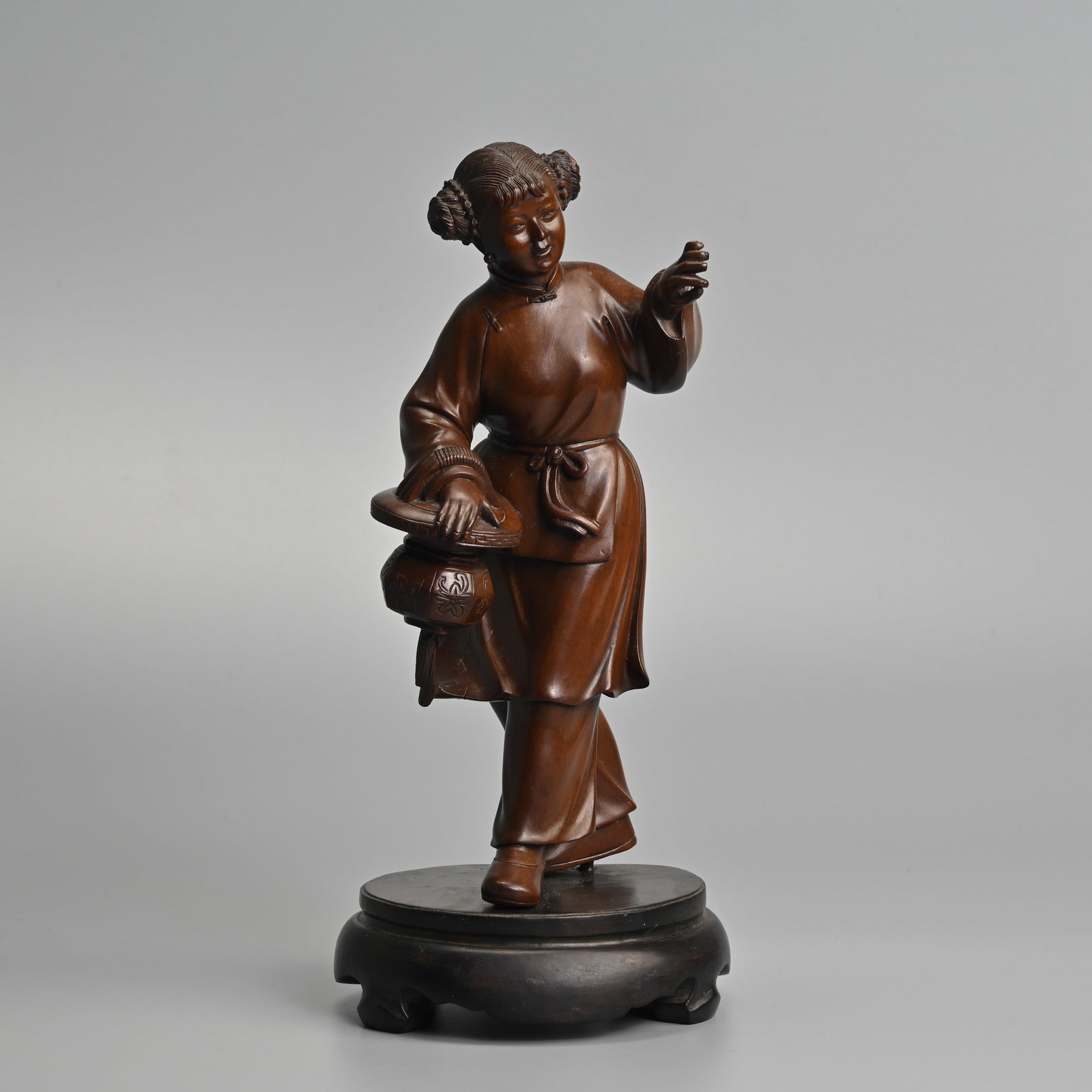 A CHINESE HARDWOOD CARVING OF A GIRL WITH BASKET, 20TH CENTURY.: Modelled in stride holding a basket over her right arm, on display stand. 24.5cm.